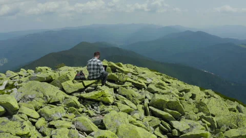 A man works on a laptop while sitting on top of a rocky mountain. Stock Footage 248205280