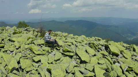 A man works on a laptop while sitting on top of a rocky mountain. Stock Footage 248205394