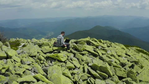A man works on a laptop while sitting on top of a rocky mountain. Stock Footage 248205456