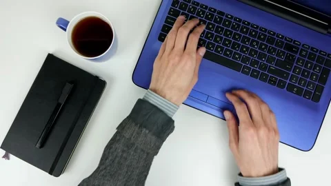 A man works with a laptop on a white table. Notepad, pen, cup of tea. Stock Footage 101752382