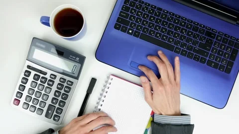 A man works with a laptop on a white table. Notepad, calculator, cup of tea Stock Footage 101753354
