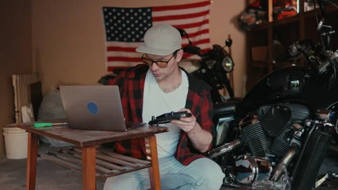 Man works on a project in a garage while sitting at a table with a laptop and Stock Footage 347985808
