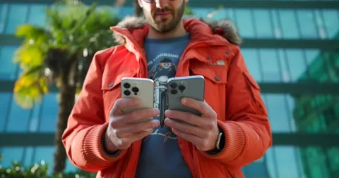 A man works on two iPhone 14 devices in front of a large glass building, Stock Footage 235908034