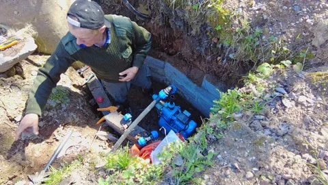 Man works on water pump system in deep hole during sunny day in rural area Stock Footage 337432004