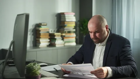 A man works at a work station at a computer, then interrupts papers with graphs. Stock Footage 222439980