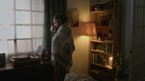 Man Wrapped in Blanket Drinking Coffee and Looking through Window at Home Stock Footage 341680847