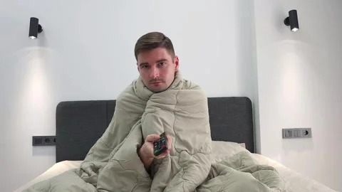 Man wrapped in blanket sitting on bed holding TV remote control. Video stock 284044809