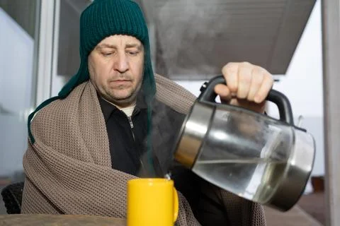 Man wrapped in a warm blanket on a chilly outdoor terrace pours hot water i.. Stock Photos