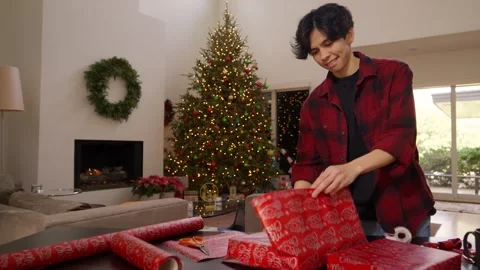 A man wraps gifts at a table in a living room decorated with a Christmas tree Stock Footage 341027183