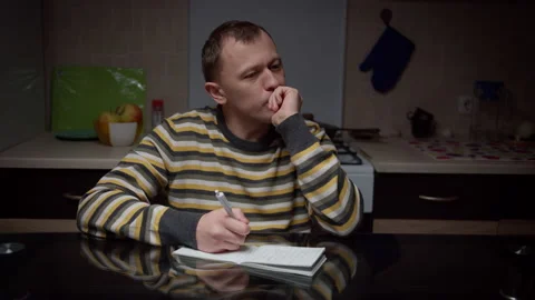 Man writes in a notebook his plans, sitting at the kitchen table in the evening Stock Footage 135304564