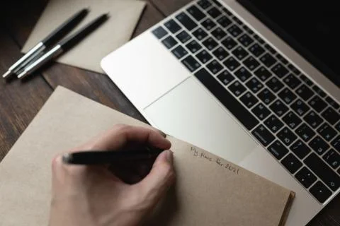 A man writes in a notebook My plans for 2021 with a pen on a dark wooden table. Stock Photos