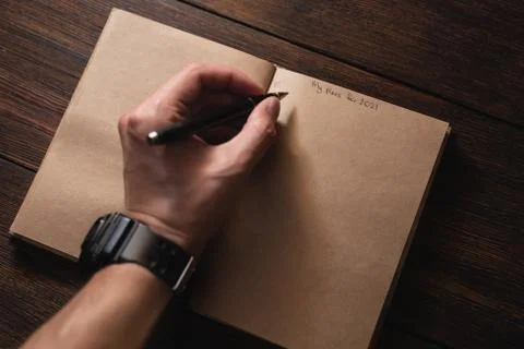 A man writes in a notebook My plans for 2021 with a pen on a dark wooden table. Stock Photos