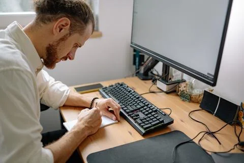 Man writes in notebook while sitting at desk next to computer in modern Stock Photos