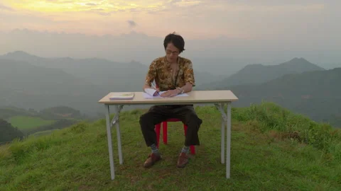 Man Writing At A Folding Desk On A Grassy Ridge, With Mountains Fading Into The Stock Footage 345278023
