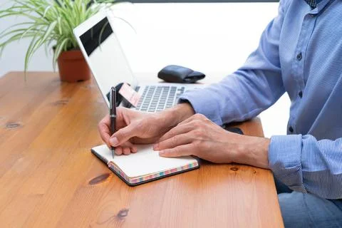 Man writing in a notebook with a computer in the background. Stock Photos