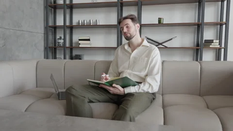 Man Writing In Notebook On Couch, Focused On Creative Thought In Stylish Home 스톡 동영상 306696994