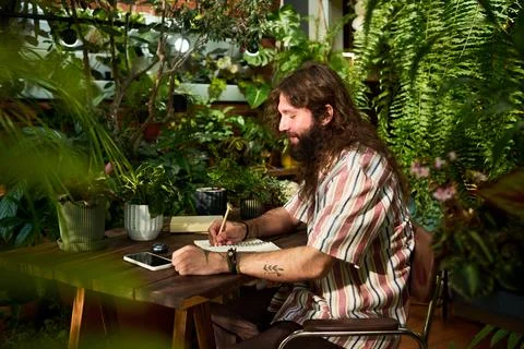 Man Writing in Notebook Surrounded by Green Plants at Home Fotos de archivo