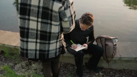 Man Writing in Notebook by Water with Another Man Holding Book featuring woman Stock Footage 348096870