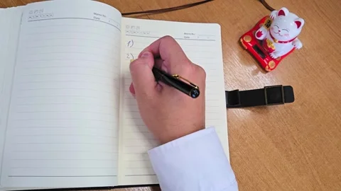 Man writing notes in planner beside Maneki neko cat figurine Stock Footage 315686652