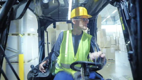 A man in a yellow hard hat and safety vest drives a forklift in a warehouse. 库存影片 307998063
