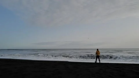 Man in a yellow jacket standing on the black sand beach overlooking the horizon Видео 257307473