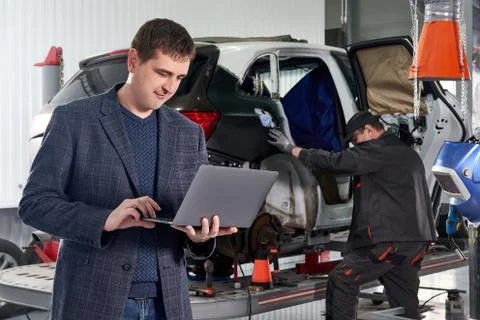 Manager holding laptop while standing near car in auto repair shop with mechanic Stock Photos