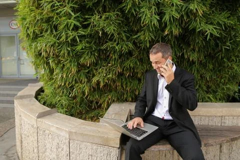 Manager talking by smartphone and working with laptop outside in Stock-Fotos