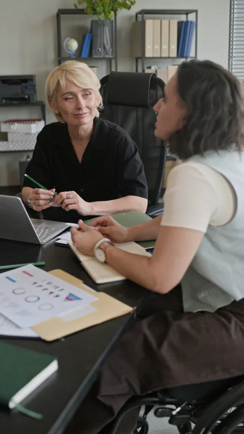 Manager in Wheelchair Talking to Colleague at Table with Laptop and Papers Stock Footage 335756928