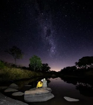 Mand and woman sit on a boulder together next to a river watching the milkway Stock Photos