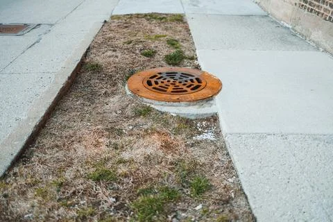 A manhole cover rests on a small patch of grass beside a concrete sidewalk. T 스톡 사진