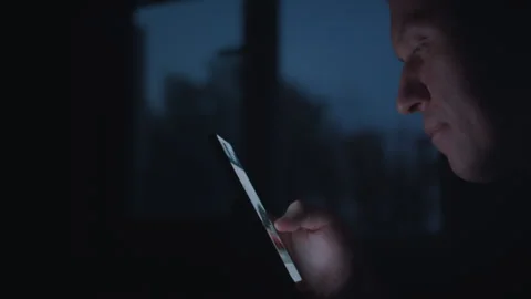 A mans face against the backdrop of a window at night, browsing apps on his Stock Footage 316954016