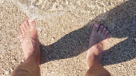 Mans feet standing in shallow clear sea water on a pebble beach Stock Footage 319418086