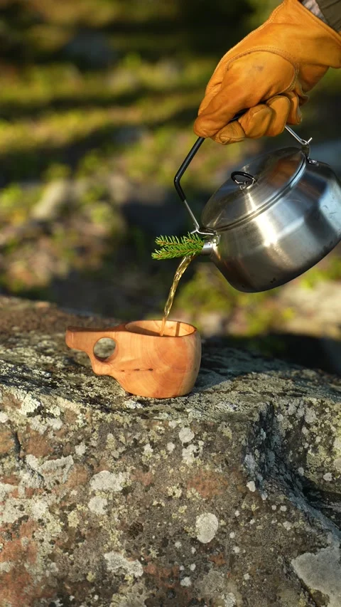A man's gloved hand is pouring tea from a camping kettle into a mug. A kuksa and Vidéo 308290033