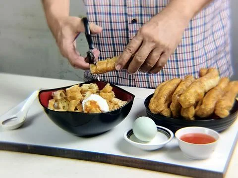 Man's hand cuts fried bread or cakwe using scissors over a bowl of porridge.. 스톡 사진