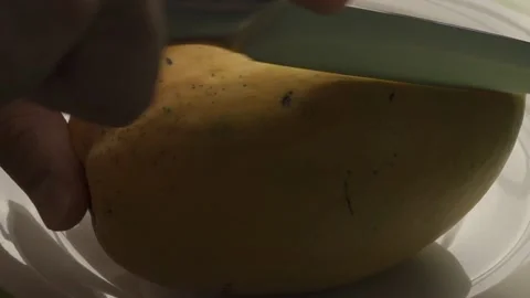 A man's hand cuts a ripe mango with a sharp knife on a white plate in close-up Stock Footage 232969359
