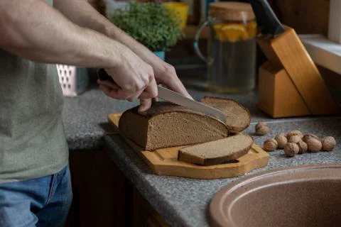 Man's hand cutting rye bread on wooden board with knife on rustic style kitchen Stock Photos