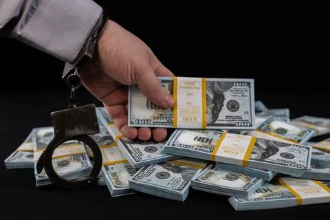 A man's hand in a handcuff holds a stack of hundred dollar bills over a pile of Stock Photos