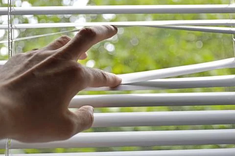 Man's hand opens strips of blinds to look through the window Stock Photos