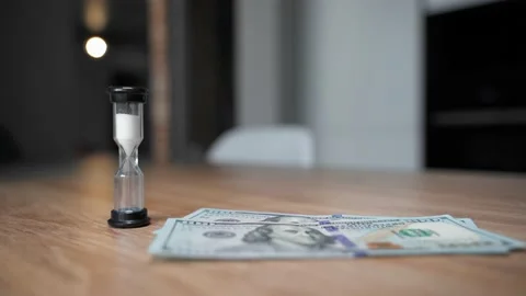 A man's hand puts an hourglass on the table. Falling 100 dollar banknotes Stock Footage 151971248
