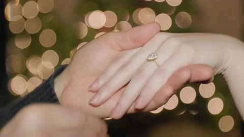Mans hand putting wedding ring on womans hand in front of Christmas lights. 4K Stock Footage 230203012