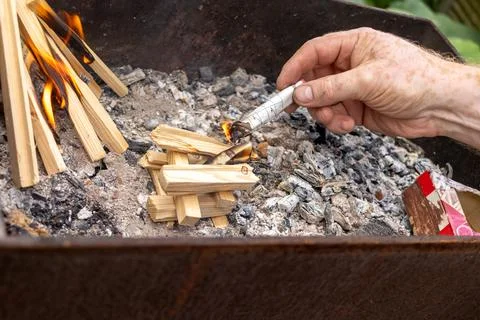 Man's hand sets fire to stacked wood with newspaper in barbecue pit, illust.. Stock Photos
