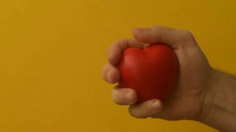 Man's hand squeezes red anti-stress heart toy on a yellow background Stock Footage 147367651