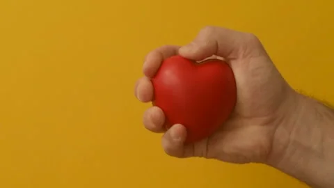 Man's hand squeezes red anti-stress heart toy on a yellow background Stock Footage 147367709