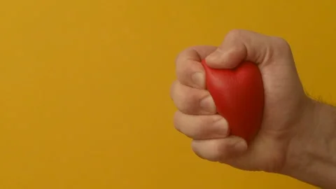 Man's hand squeezes red anti-stress heart toy on a yellow background Stock Footage 147367773