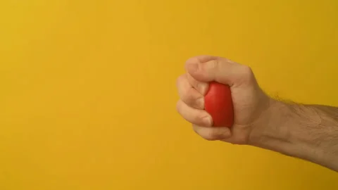 Man's hand squeezes red anti-stress heart toy on a yellow background Stock Footage 147367859
