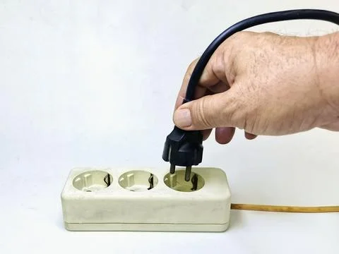 A man's hand tries to plug a black electrical cord into an electrical outle.. 스톡 사진