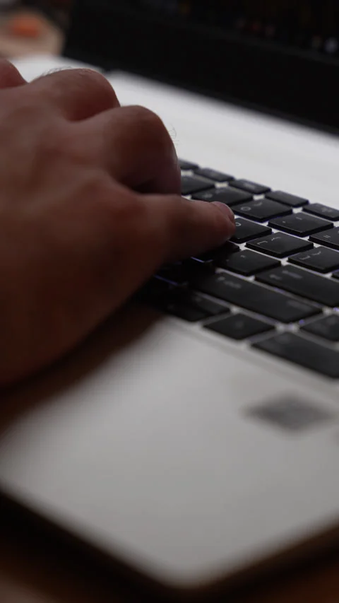 A man’s hand types on the black keyboard of a silver laptop in a dim room, the Stock Footage 347743796