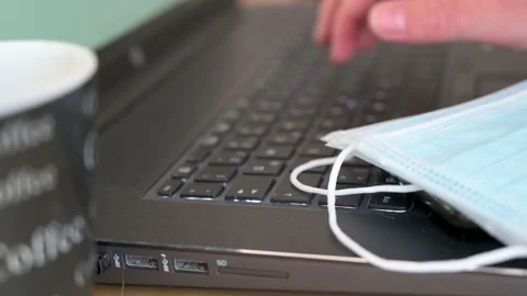 Man's hand is typing on a laptop keyboard next to a face mask to protect aga Stock Footage 132281973