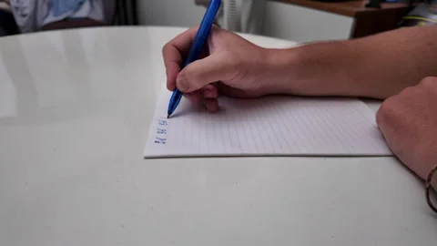 A man's hand writes text with a blue pen on a sheet of notebook paper. Stock-Footage 332203457
