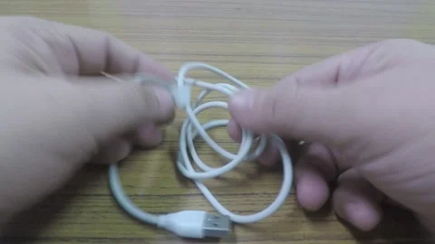 MAN'S HANDS UNTANGLING A WHITE USB CABLE ON TOP OF A WOODEN TABLE Stock Footage 133634430